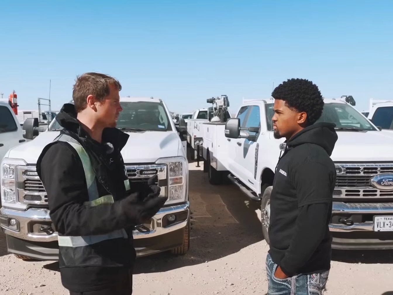 Two men in jackets stand facing each other in a truck lot with white pickup trucks